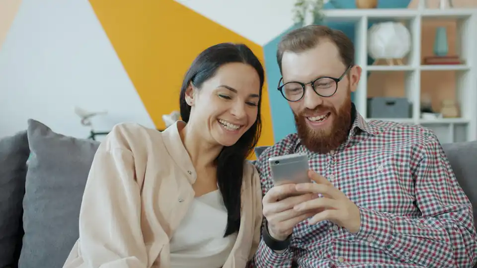 Casal sorrindo no sofá enquanto olha o aplicativo do banco no celular