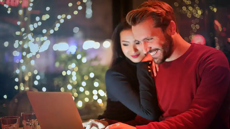 Casal sorrindo em frente a um notebook com luzes de Natal desfocadas ao fundo